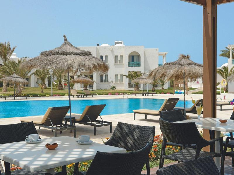 Terasa s stoly a židlemi u bazénu, slámové slunečníky a bílá budova v pozadí.