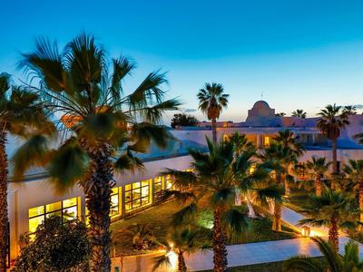 Palmen umgeben ein modernes Hotelgebäude bei Sonnenuntergang mit blauem Himmel und warmem Licht.