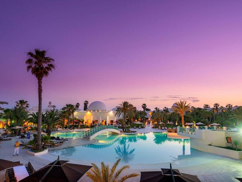 Großer Pool mit Brücke, umgeben von Palmen und beleuchteten Liegen bei violettem Abendhimmel.