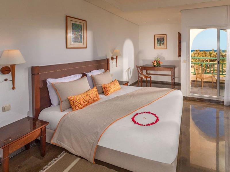 Helles Schlafzimmer mit großem Bett, Holzmöbeln, Teppich und Balkon mit Meerblick.