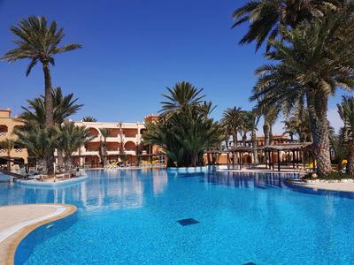 Großer blauer Pool vor einem Hotel mit Palmen und blauem Himmel im Hintergrund.