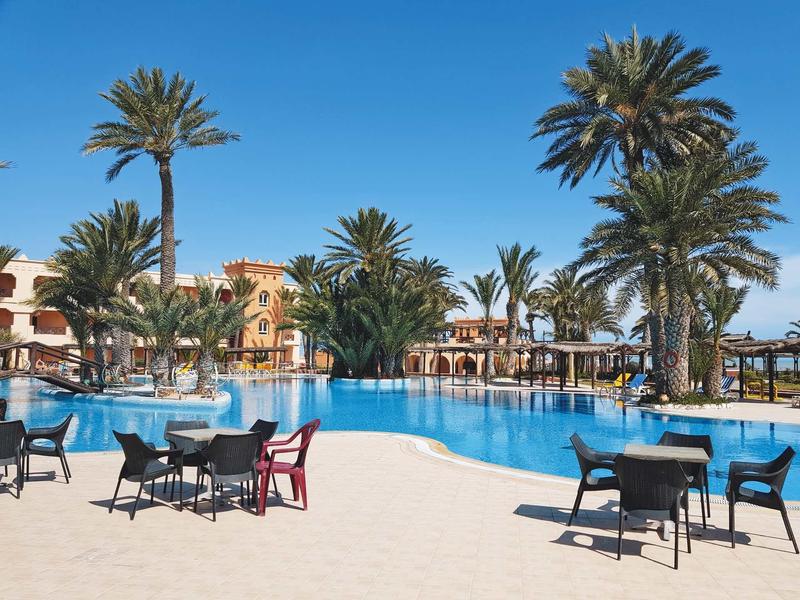 Grande piscina con palme e vari tavoli e sedie in un resort soleggiato.