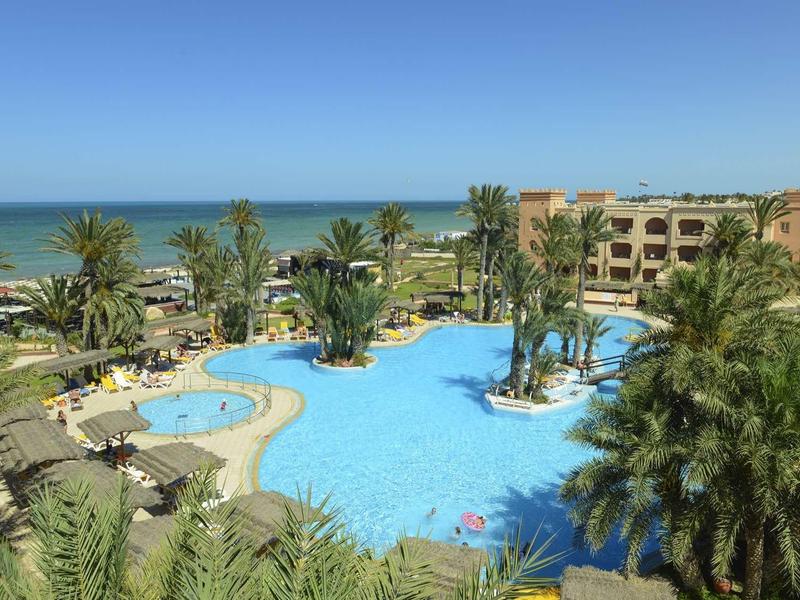 Grande piscina dell'hotel con palme e vista sul mare sotto un cielo limpido.