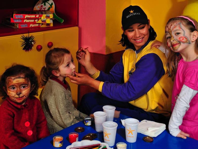 Bambini con pittura sul viso e animatore seduti a un tavolo con materiale per lavori manuali.