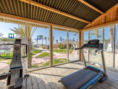 Fitnessraum mit Laufband und Rudergerät mit Blick auf Strand und Meer durch große Fenster.