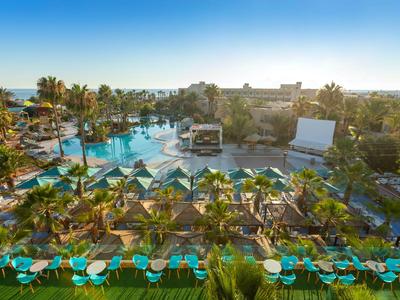 Großer Poolbereich mit Liegestühlen und Palmen in einem Resort bei Sonnenschein.