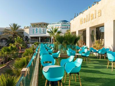 Hotelterrasse mit blauen Stühlen und Tischen, umgeben von Palmen und Klippen im Hintergrund.