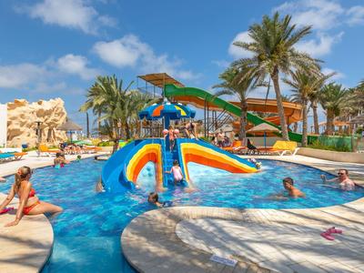 Hotelpool mit Wasserrutschen, Sonnenschirmen und Palmen unter blauem Himmel.