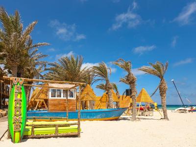 Sonniger Strand mit buntem Boot, Palmen und Holzhütten unter blauem Himmel.