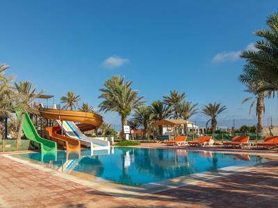 Grande piscina dell'hotel con scivoli d'acqua, circondata da palme e lettini sotto un cielo azzurro.