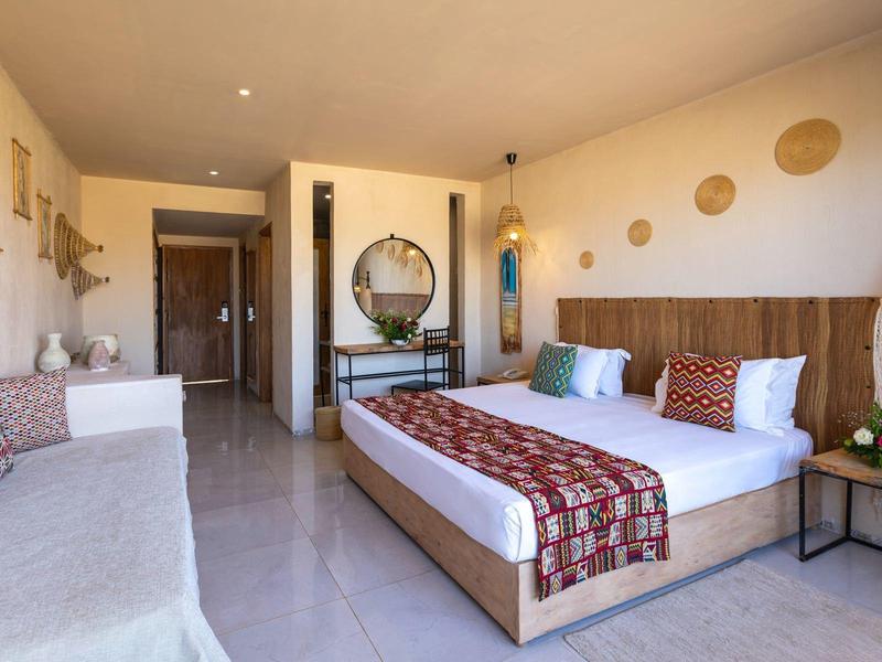 Camera d'hotel luminosa con grande letto matrimoniale, divano e arredamento moderno in toni caldi.