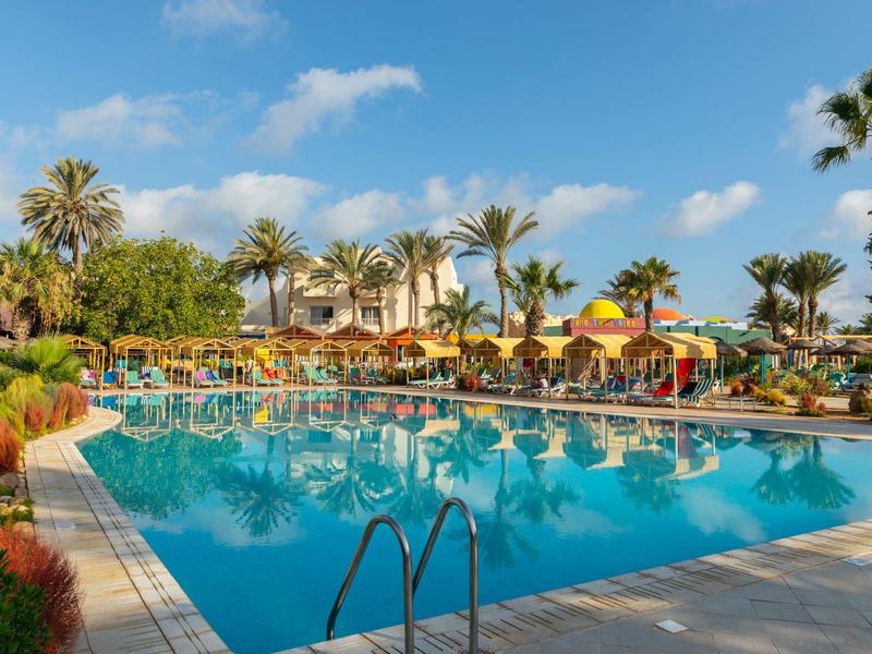 Grande piscina con lettini e palme in un complesso alberghiero sotto un cielo sereno.