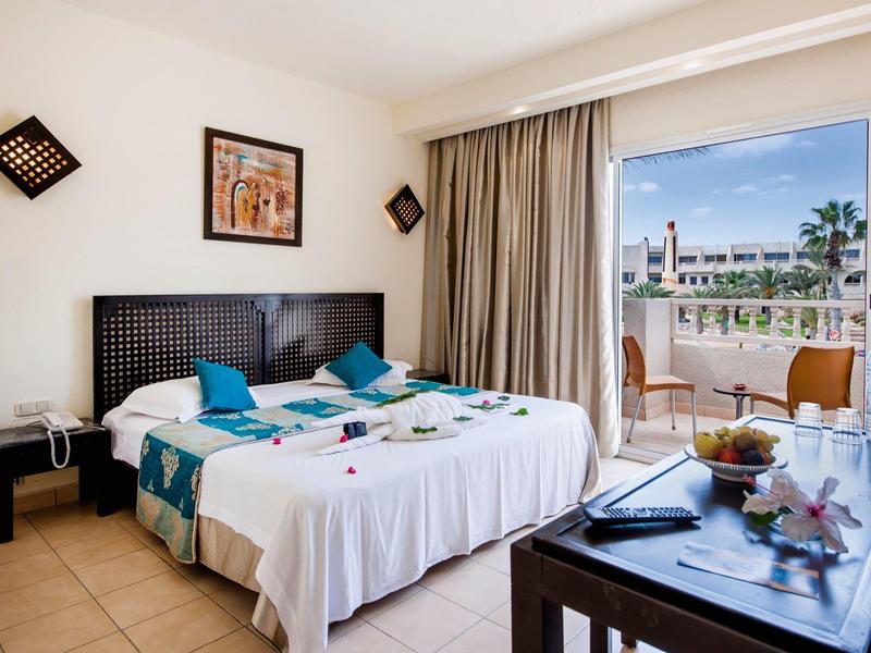 Camera d'hotel luminosa con letto matrimoniale, balcone e vista su palme e mare.