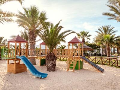 Kinderspielplatz mit Rutschen und Klettergerüsten, umgeben von Sand und Palmen bei Tageslicht.