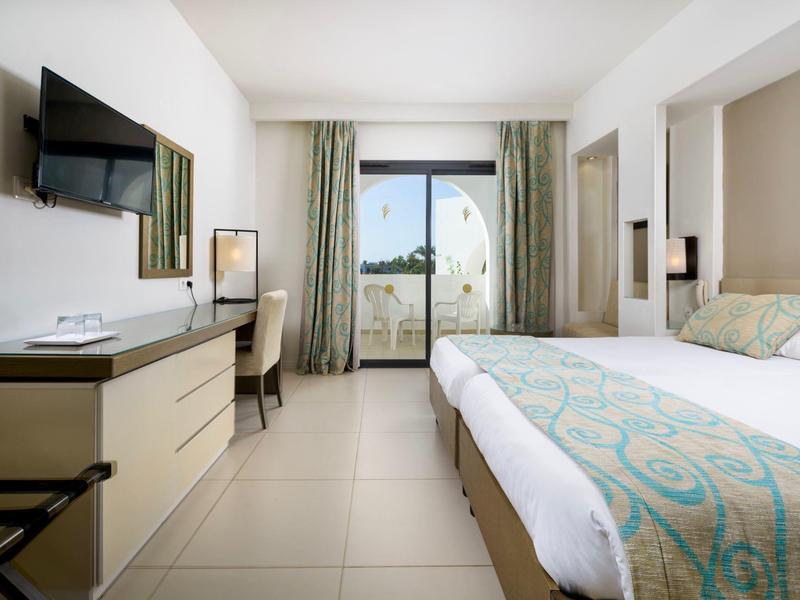 Hell eingerichtetes Hotelzimmer mit Doppelbett, Schreibtisch, Fernseher und Balkon mit kurzem Vorhang.