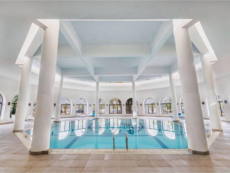 Großes Hallenbad mit weißen Säulen, glattem Boden und hohen Decken in hellem Raum.