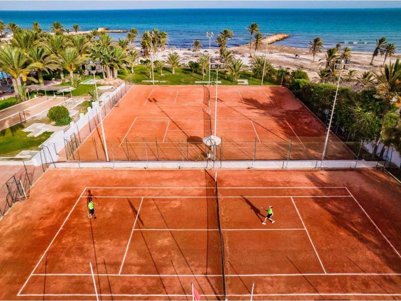 Zwei rote Sand-Tennisplätze mit Spielern, umgeben von Palmen und Blick aufs blaue Meer.