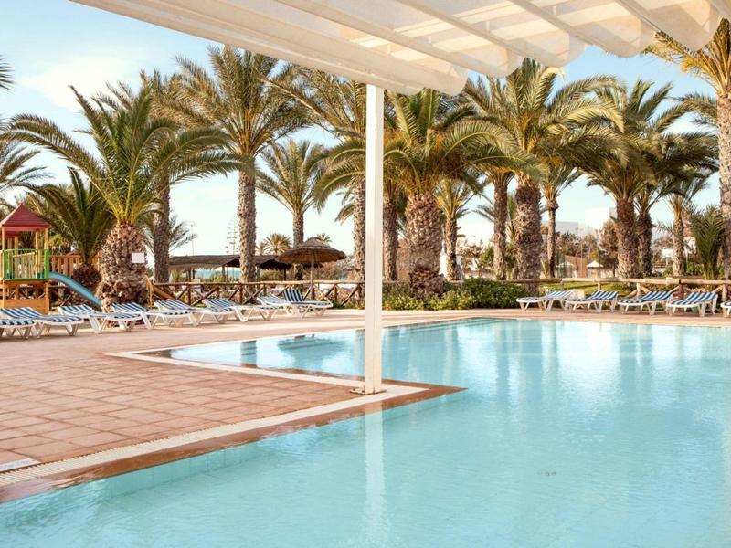 Großer Pool mit blauem Wasser, umgeben von Palmen und Sonnenschirmen unter hellblauem Himmel.