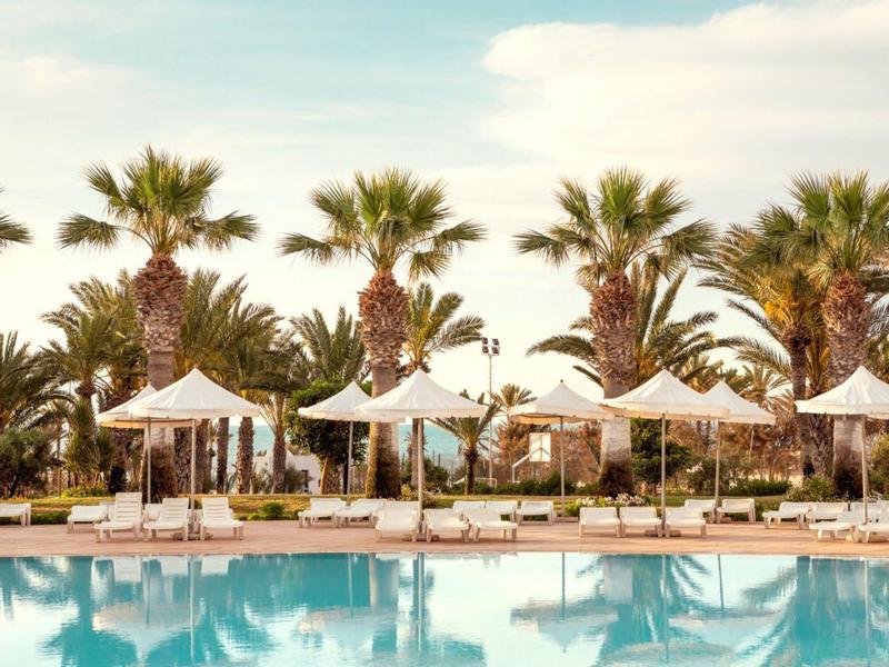 Pool mit Liegestühlen, weißen Sonnenschirmen und Palmen unter blauem Himmel.