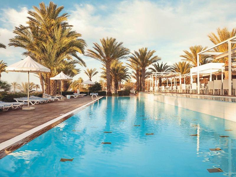 Großer Pool mit klar blauem Wasser, Palmen, Sonnenschirmen und Liegestühlen bei klarem Himmel.