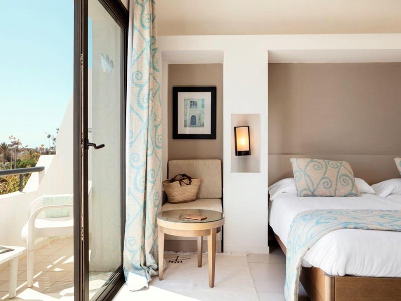 Helles Schlafzimmer mit Doppelbett, offenem Balkon und hellen Vorhängen im Tageslicht.