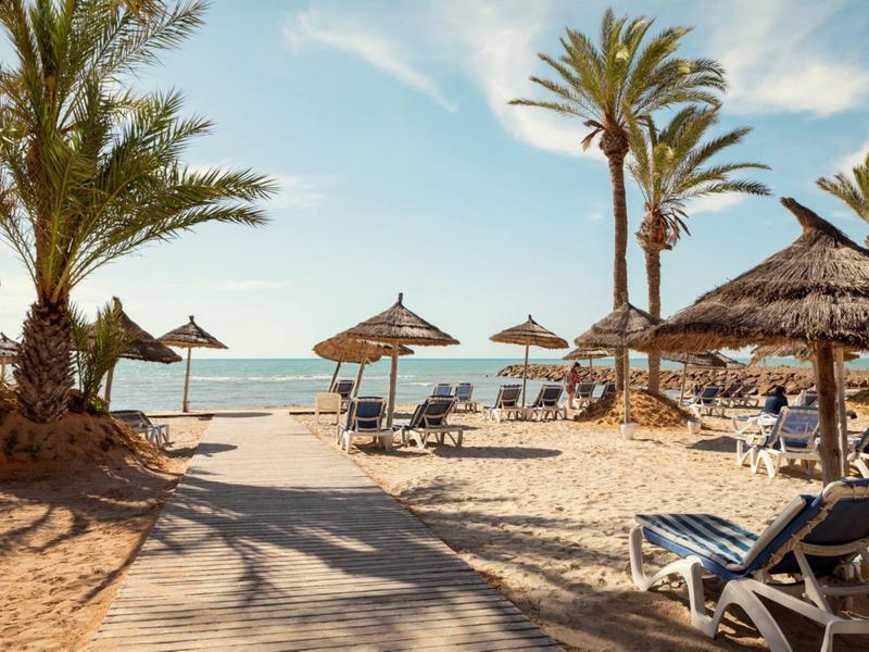 Sonniger Strand mit Palmen, Sonnenliegen und Strohhüte vor ruhigem Meer.