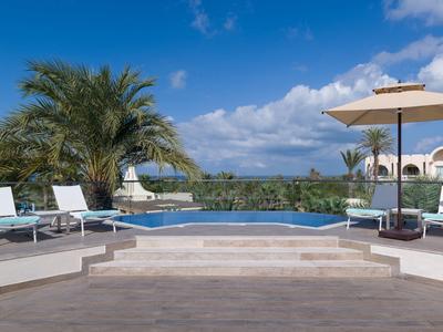 Pool mit Terrasse, Sonnenliegen und Palmen vor blauem Himmel und weißen Gebäuden.