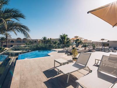Eine sonnige Terrasse mit Pool, Liegestühlen und Sonnenschirmen neben Palmen und blauem Himmel.