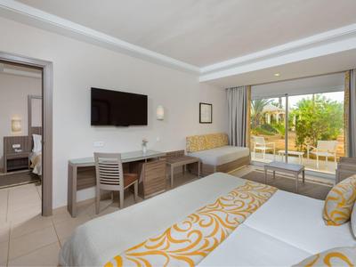 Helles Hotelzimmer mit zwei Betten, TV, Schreibtisch, Sofa und Zugang zu einer Terrasse mit grünen Pflanzen.