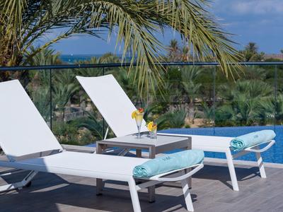 Zwei weiße Liegestühle mit blauen Handtüchern auf einer Terrasse unter Palmen.