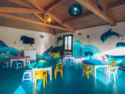 Kindergartenraum mit bunten Delfin-Wandmalereien, kleinen Tischen, Stühlen und Holzbalkendecke.