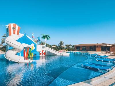 Großer Pool mit Wasserrutschen, buntem Spielbereich und blauem Himmel, Liegen am Beckenrand.