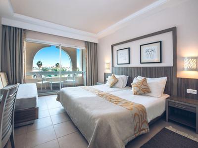 Camera d'hotel moderna con letto matrimoniale, balcone e vista sulla piscina e palme.