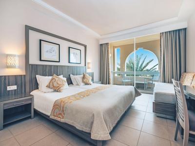 Modernes Hotelzimmer mit Doppelbett, dunkelgrauen Möbeln, Balkon und Blick auf Palmen.