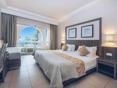 Helles Hotelzimmer mit großem Bett, Fenster mit Blick auf Meer, zwei Nachttischen und Schreibtisch.