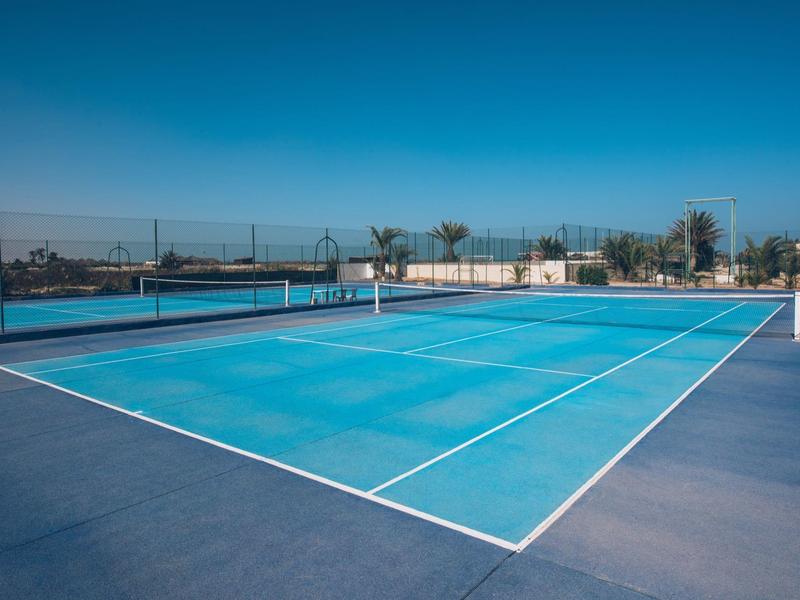 Campo da tennis vuoto con superficie blu sotto un cielo limpido in un complesso alberghiero.