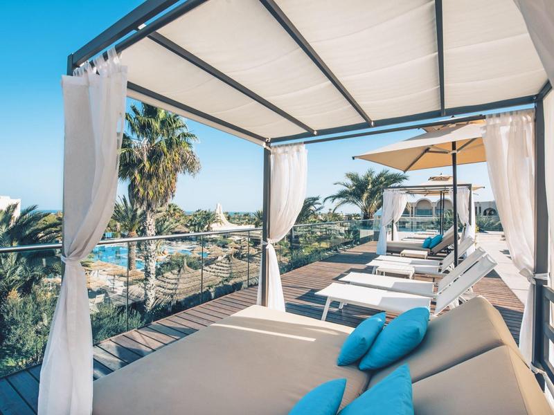 Terrazza moderna dell'hotel con lettini, tende bianche e vista sulle palme.