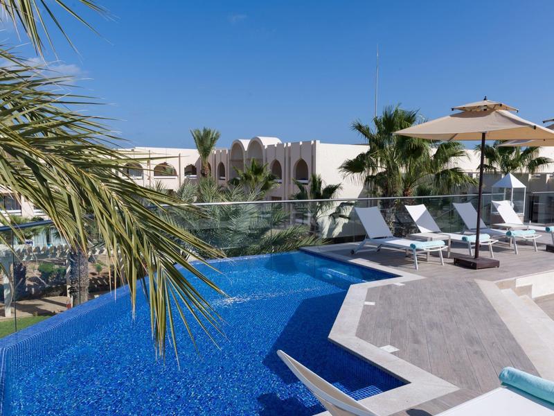 Area piscina moderna con acqua blu, lettini e palme in un resort alberghiero soleggiato.