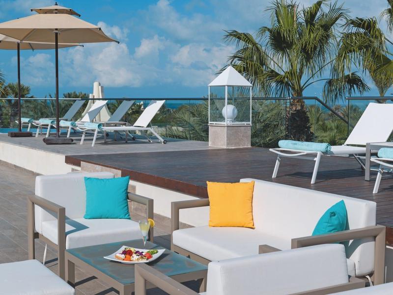 Area piscina moderna con lettini, ombrelloni e divano con cuscini colorati sotto un cielo azzurro brillante.