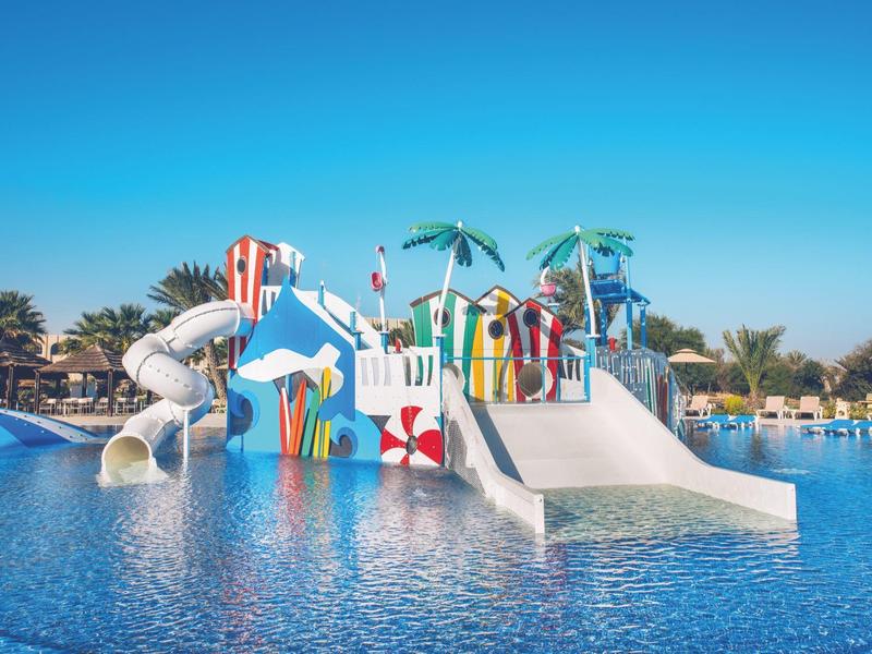 Area giochi acquatica colorata con scivoli e palme in una grande piscina sotto un cielo sereno.