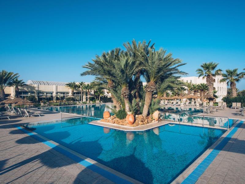Grande piscina dell'hotel con isola di palme e numerose sdraio sotto un cielo azzurro.