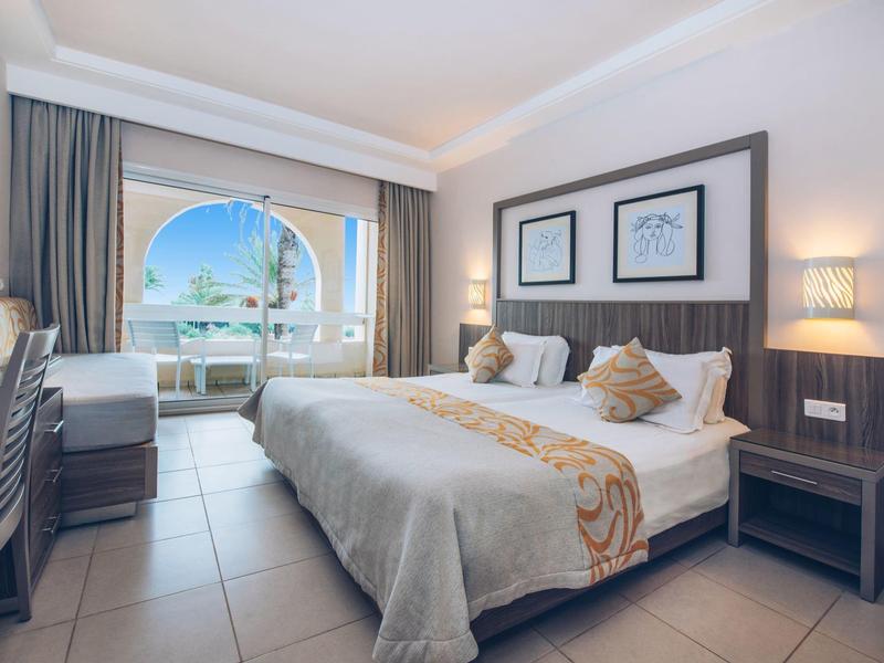 Camera d'hotel luminosa con letto matrimoniale e vista su terrazza e palme.