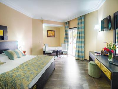 Helles Hotelzimmer mit großem Bett, Schreibtisch, Fernseher und zwei Fenster mit blauen Vorhängen.