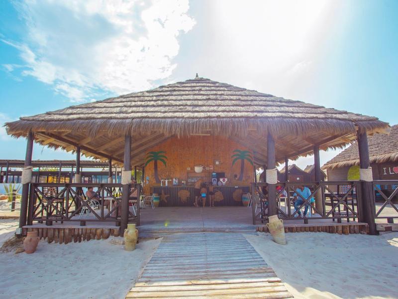 Struttura da spiaggia aperta con tetto di paglia e mobili in legno circondata da sabbia e cielo sereno.
