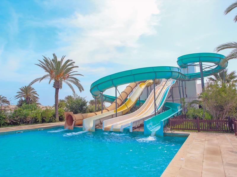 Scivolo d'acqua a più corsie vicino a una piscina con palme e cielo azzurro sullo sfondo.