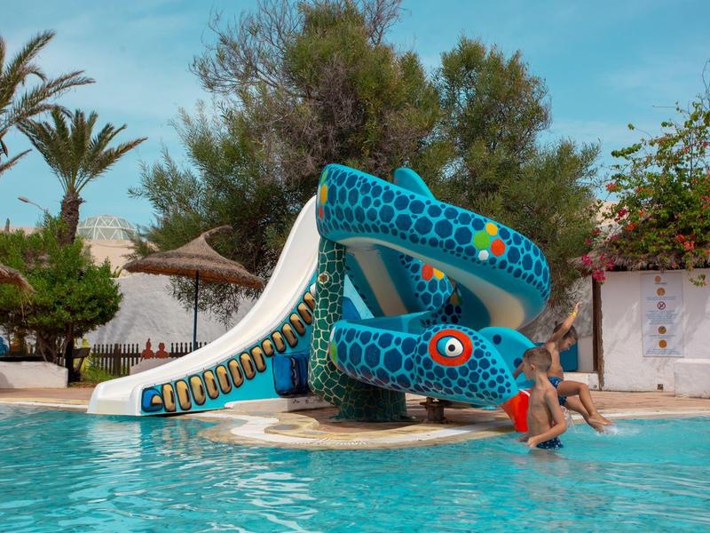 Scivolo d'acqua a forma di drago colorato in piscina con persona, palme e ombrelloni sullo sfondo.