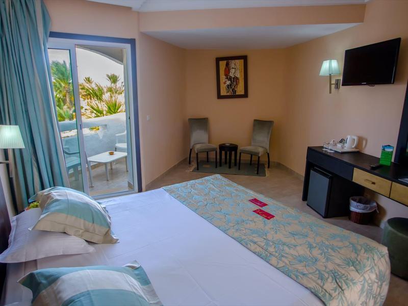 Camera d'hotel luminosa con letto matrimoniale, sedie e balcone con vista sulle palme.