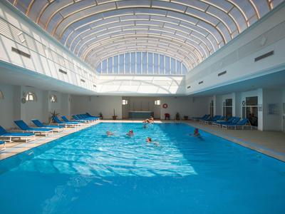 Großes Hallenbad mit blauer Schwimmfläche, Sonnenlicht und blauen Liegestühlen am Rand.