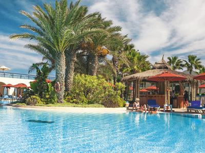 Großer Pool mit klar blauem Wasser, Palmen, Liegestühlen und Sonnenschirmen unter blauem Himmel.