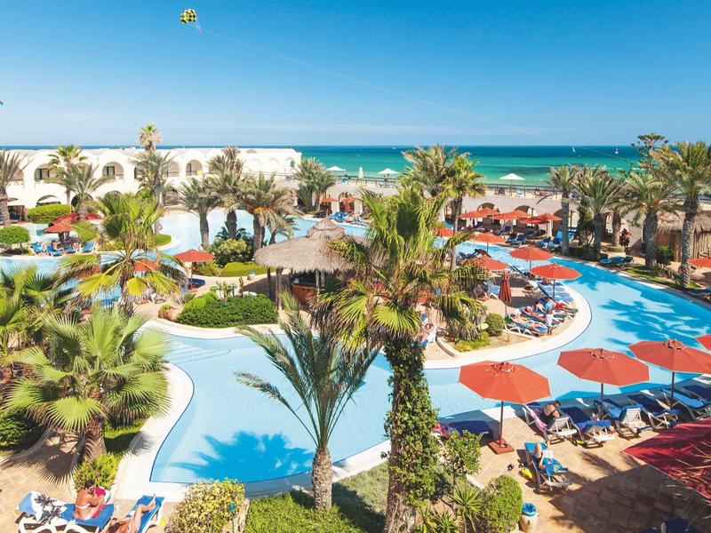 Piscina dell'hotel con palme, lettini e ombrelloni rossi vicino al mare sotto un cielo sereno